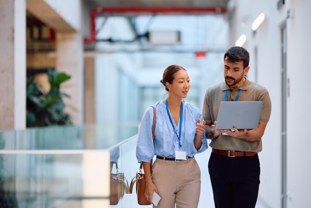 Two coworkers walk together down an open hallway, sharing a discussion while one holds an open laptop.