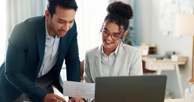 Two business people smile and collaborate over a laptop and a printout of data.