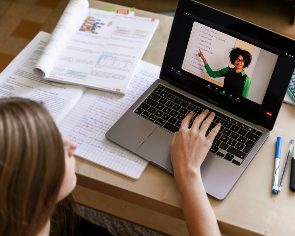 A top-down photo of a young woman participating in an online lecture from her laptop. Her hand hovers over the keyboard, and a work book and notebook are strewn across the tabletop.