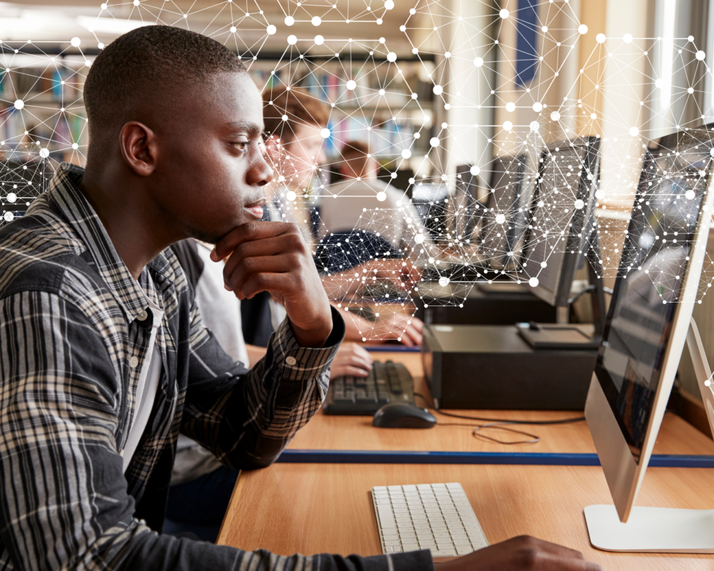 A man sits in front of a computer monitor at the end of a table lined with other monitors and users. He studies the screen intently, hand resting on his chin. There is an overlay of dots and lines forming a hexagonal network of shapes overlaid above the image.