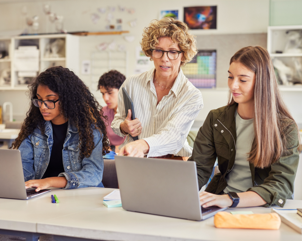 Two female college students work on computers at a shared desk. A professor reaches between them, gesturing to one student's laptop screen.