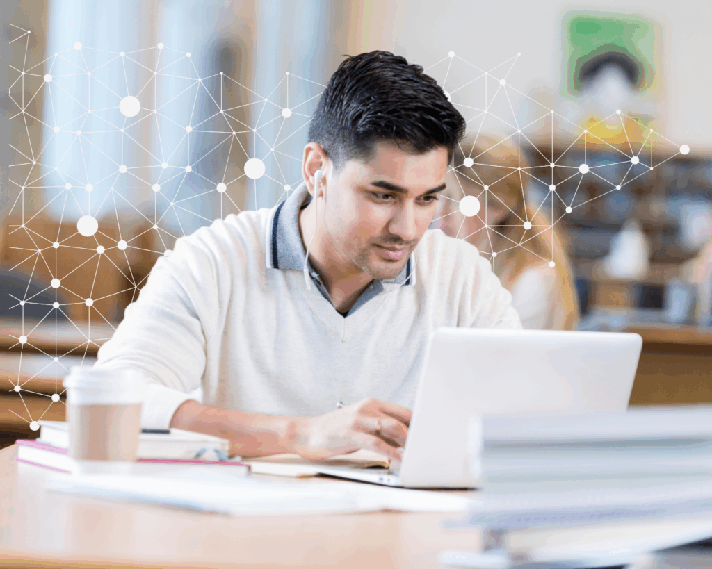 A photo of a male student working on a laptop in a library. He has earbuds in, and a to-go coffee next to him.
